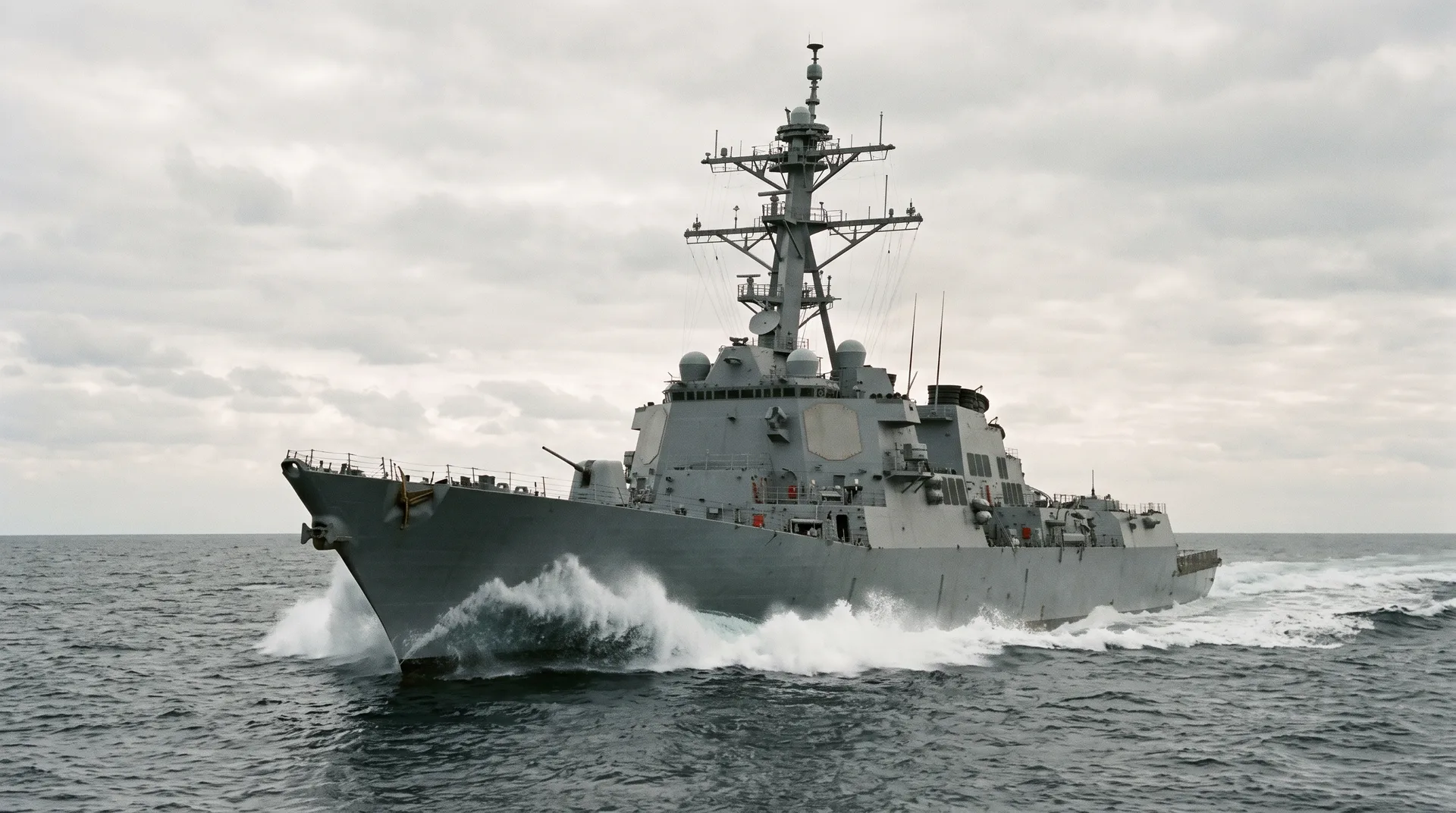 A grey naval warship under way at sea, seen from ahead and to one side, its angular superstructure rising above a breaking bow wave.