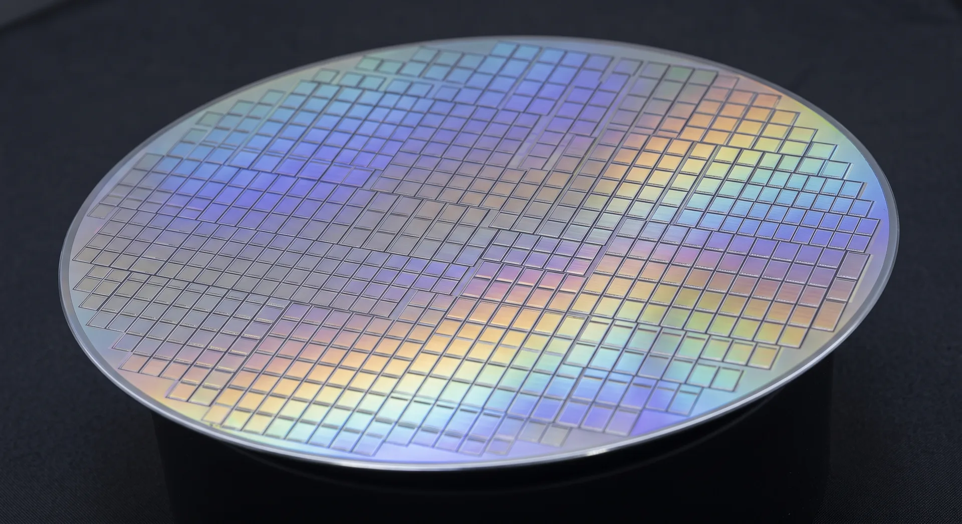 A mirror-bright circular silicon wafer on a stand, its surface divided into a fine grid of identical chips and throwing blue and gold iridescence.
