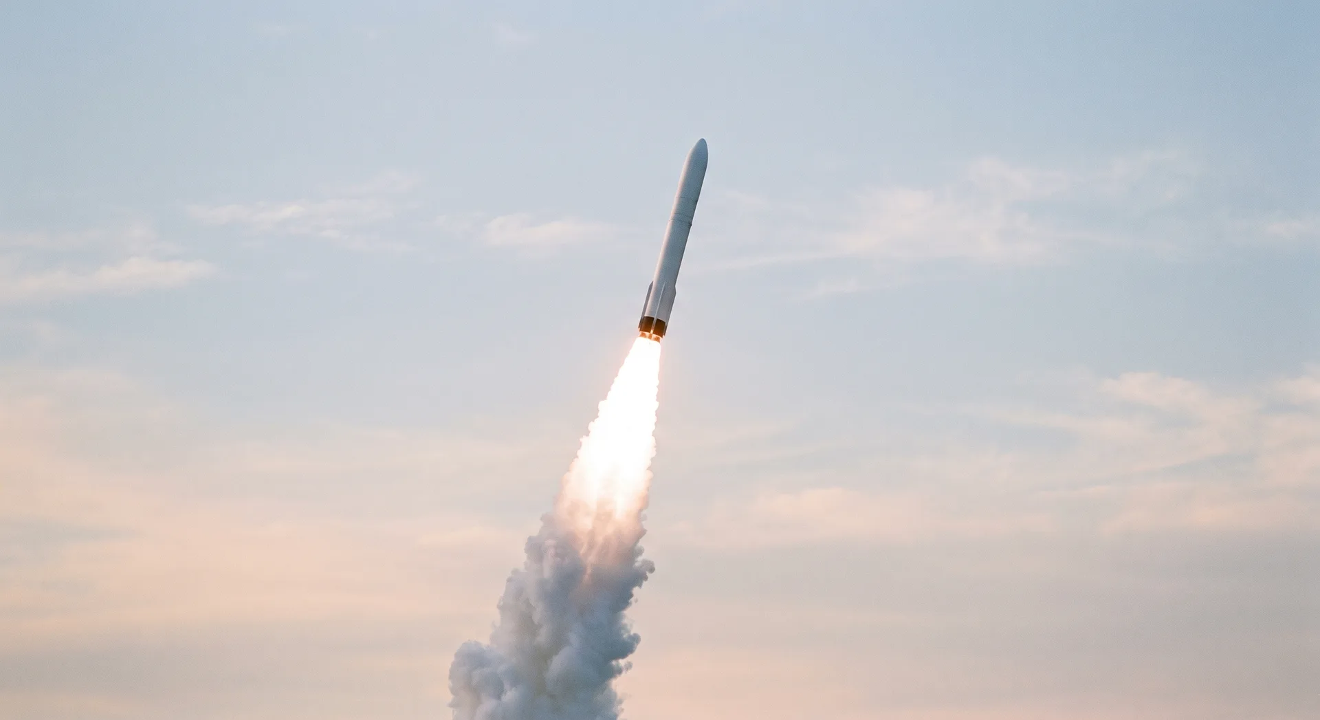 A white rocket climbing steeply against a pale morning sky, a brilliant exhaust plume trailing beneath it and smoke spreading far below.