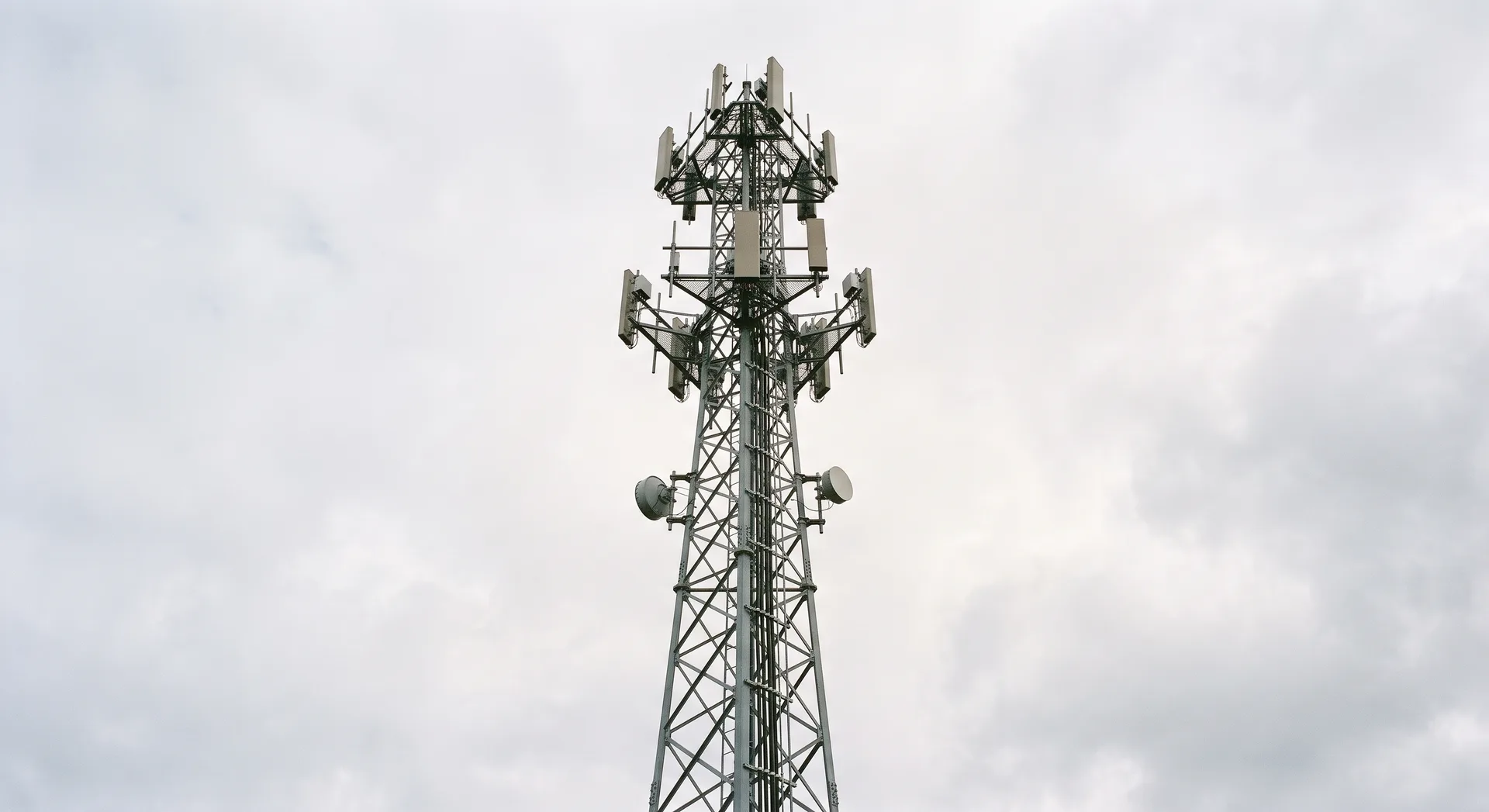 A tall lattice mobile phone mast seen from below against pale sky, with flat panel antennas and small round dishes mounted in tiers near the top.