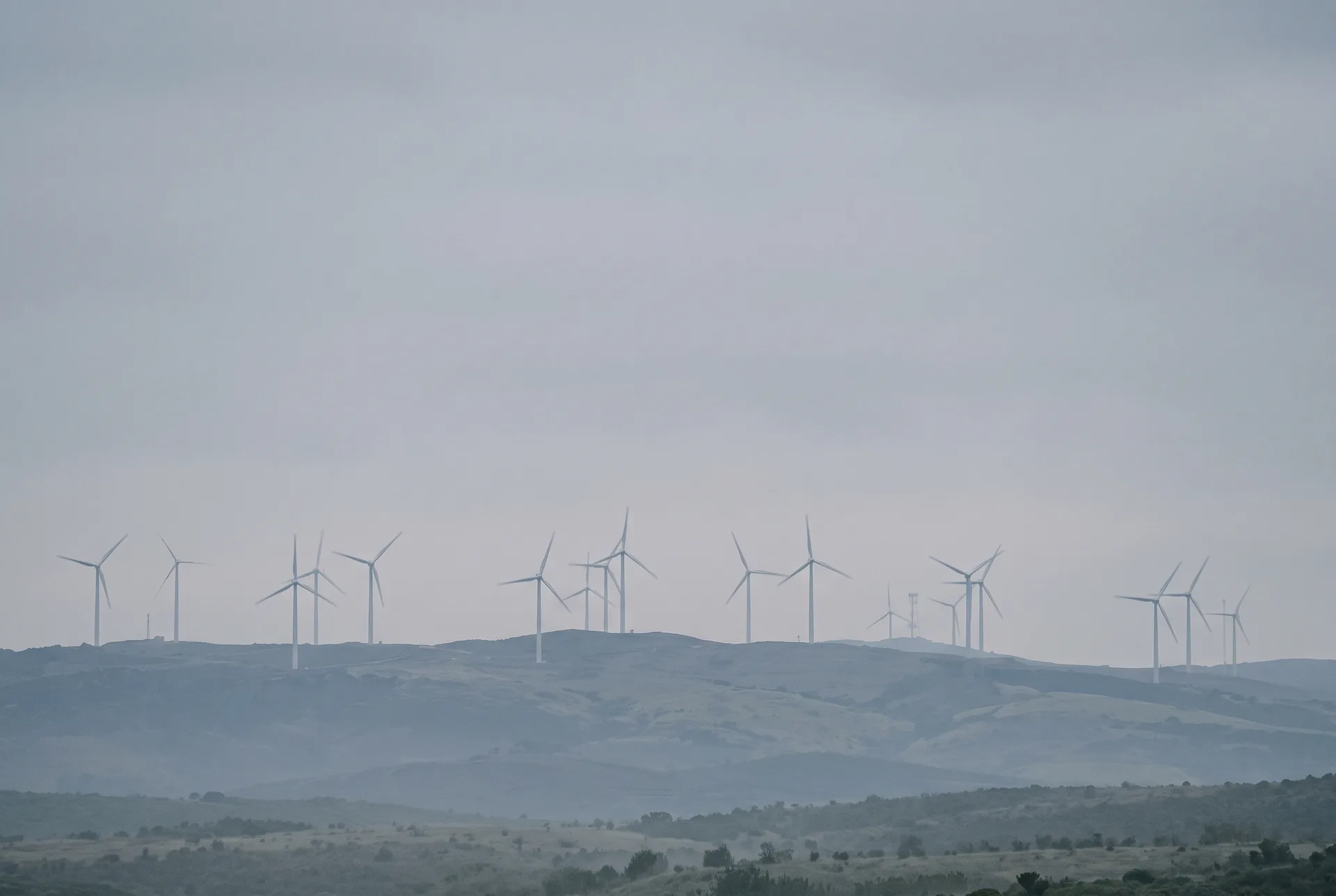 Rows of wind turbines along a hazy ridgeline, pale against a grey sky, with rolling hills receding below them.
