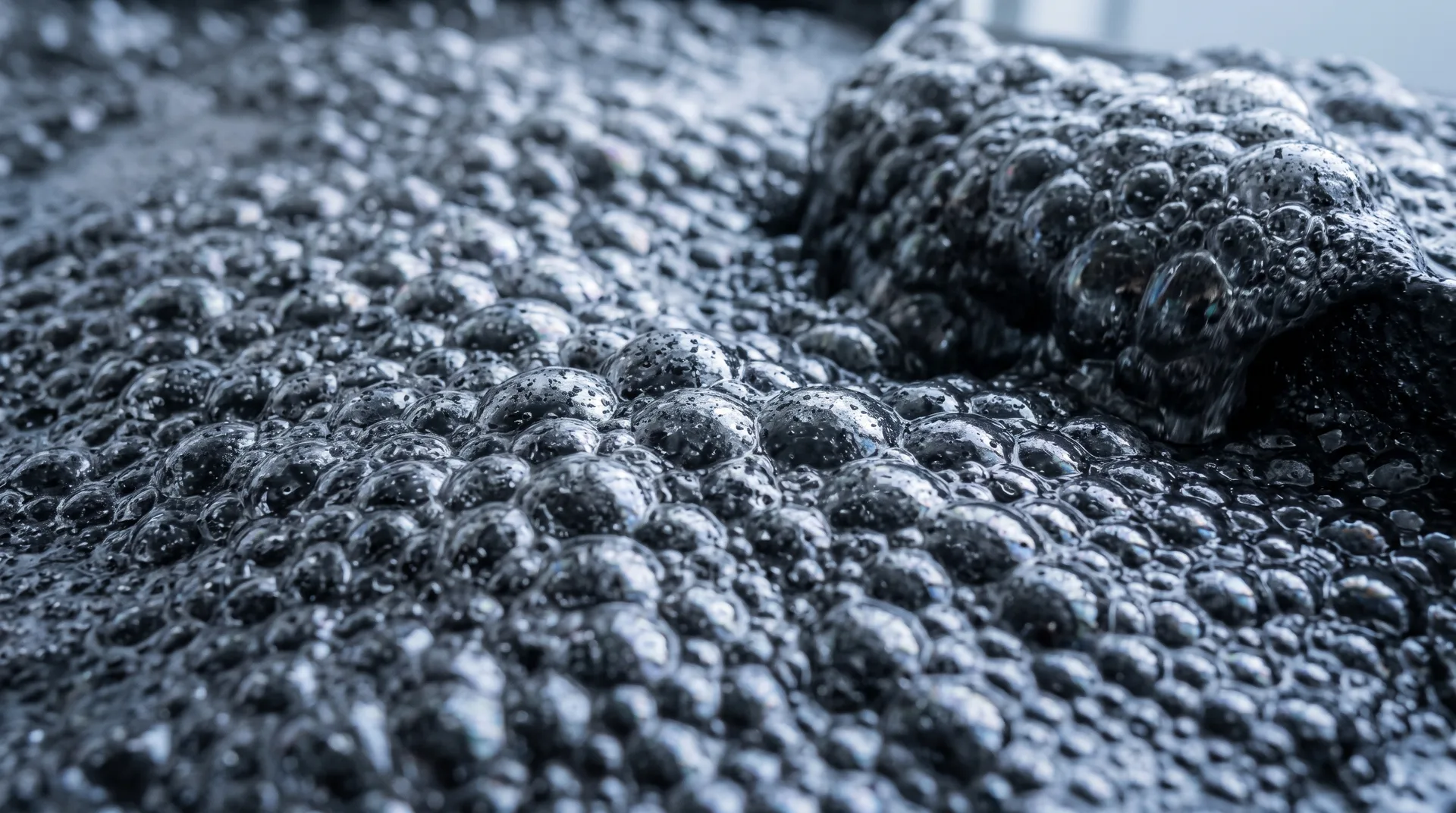 A close view of dense dark froth, wet bubbles crowded across the surface with fine particles clinging to their skins.