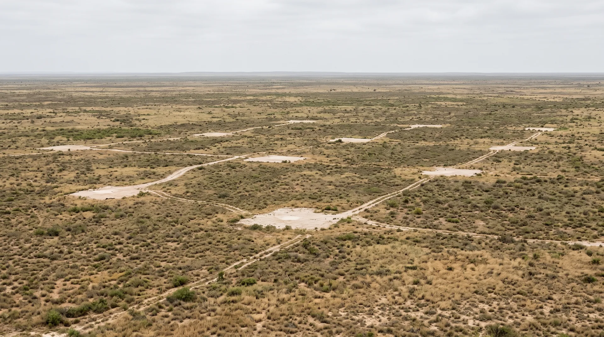 Flat dry scrubland running to a hazy skyline, with small pale cleared pads scattered across the middle distance linked by faint wheel tracks.