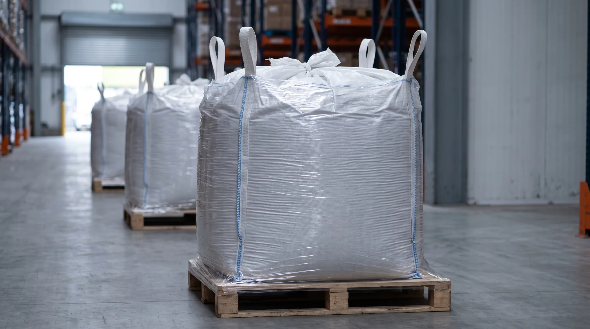 A sealed woven bulk bag standing filled and closed on a pallet in a warehouse aisle, two identical bags on pallets receding behind it.