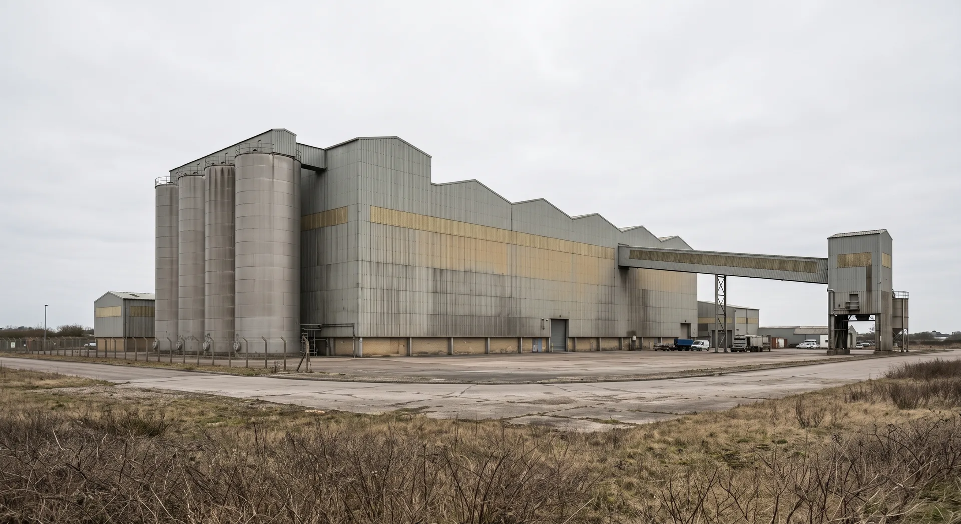 A long clad process building with four tall silos at one end and an enclosed conveyor gantry running out to a transfer tower.