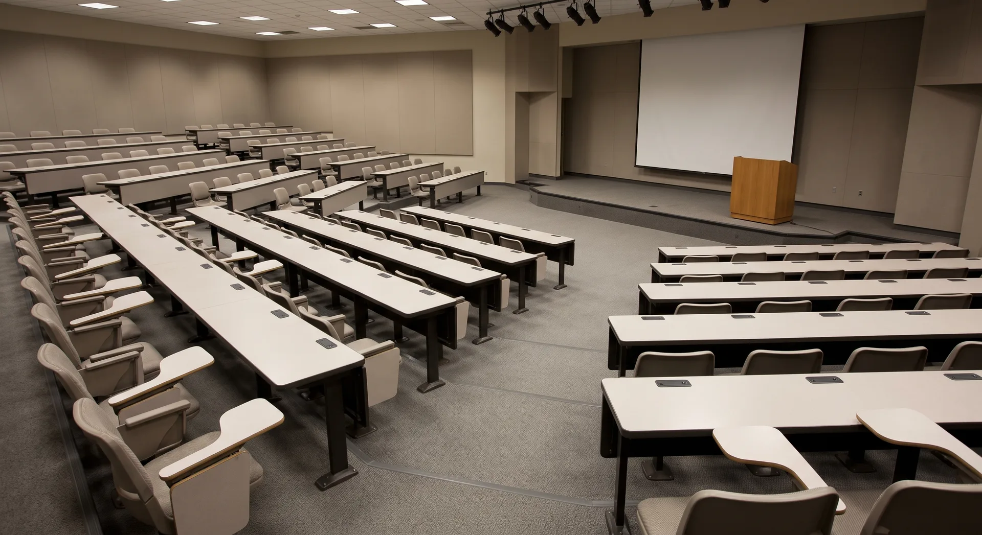 Rows of empty tiered seating running down toward a low stage with a plain lectern to one side and a blank screen behind it.