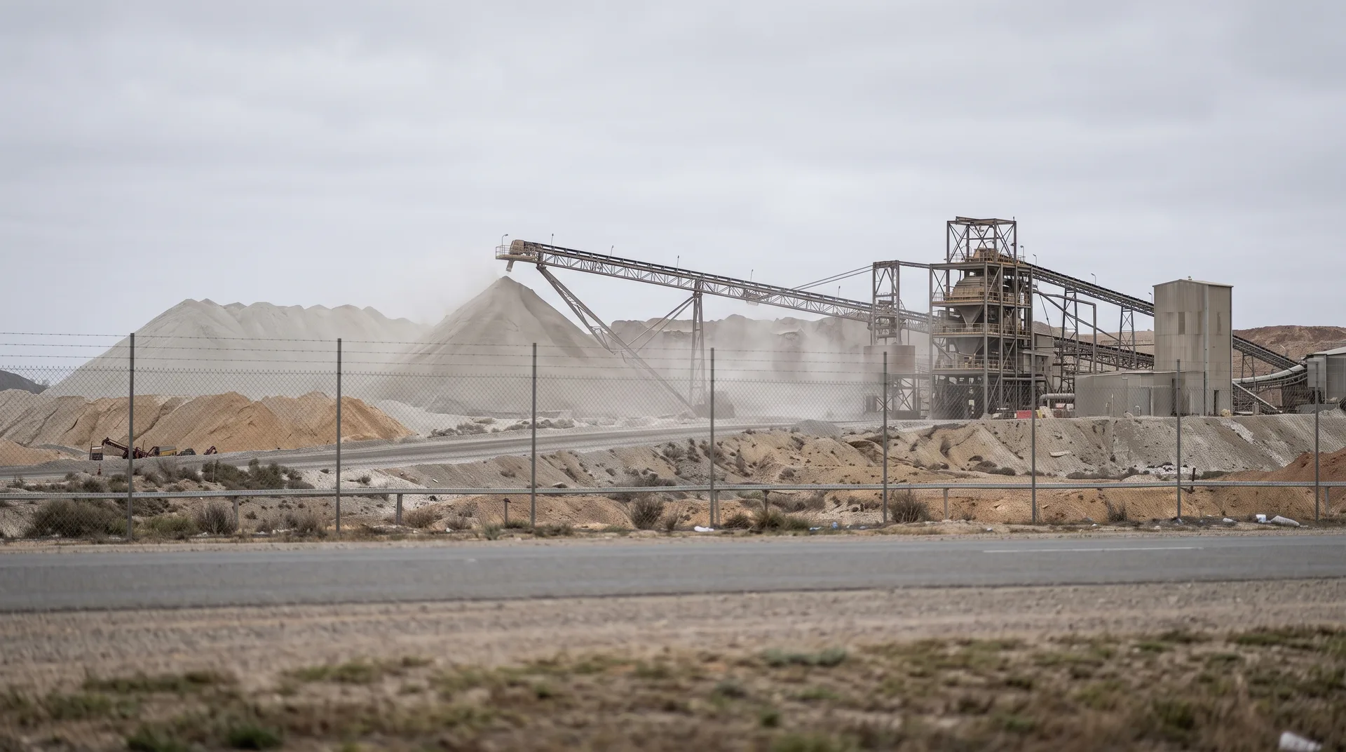 A wire perimeter fence across the foreground with a working site beyond it — pale stockpiles, a plant structure and dust over a haul road.