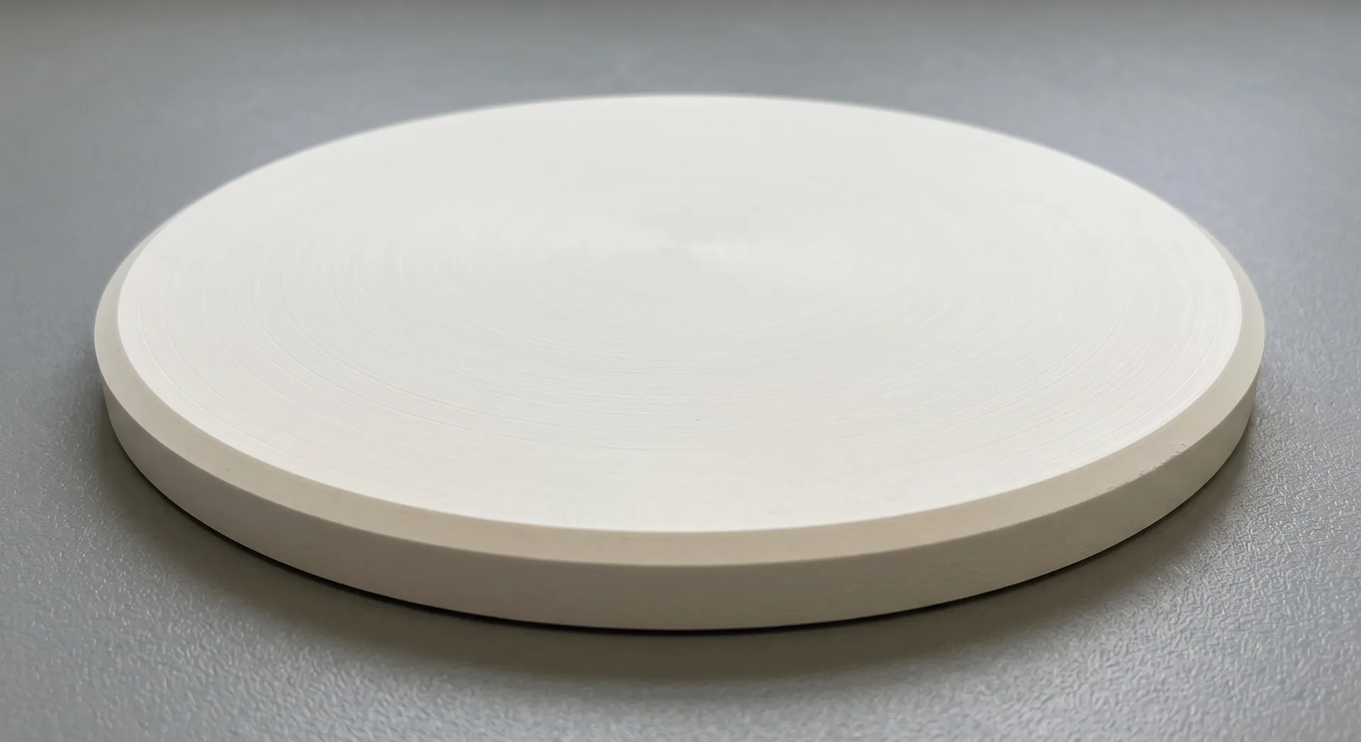 A round off-white ceramic disc lying flat on a grey surface, seen at a low angle so its smooth face and plain machined edge both read.