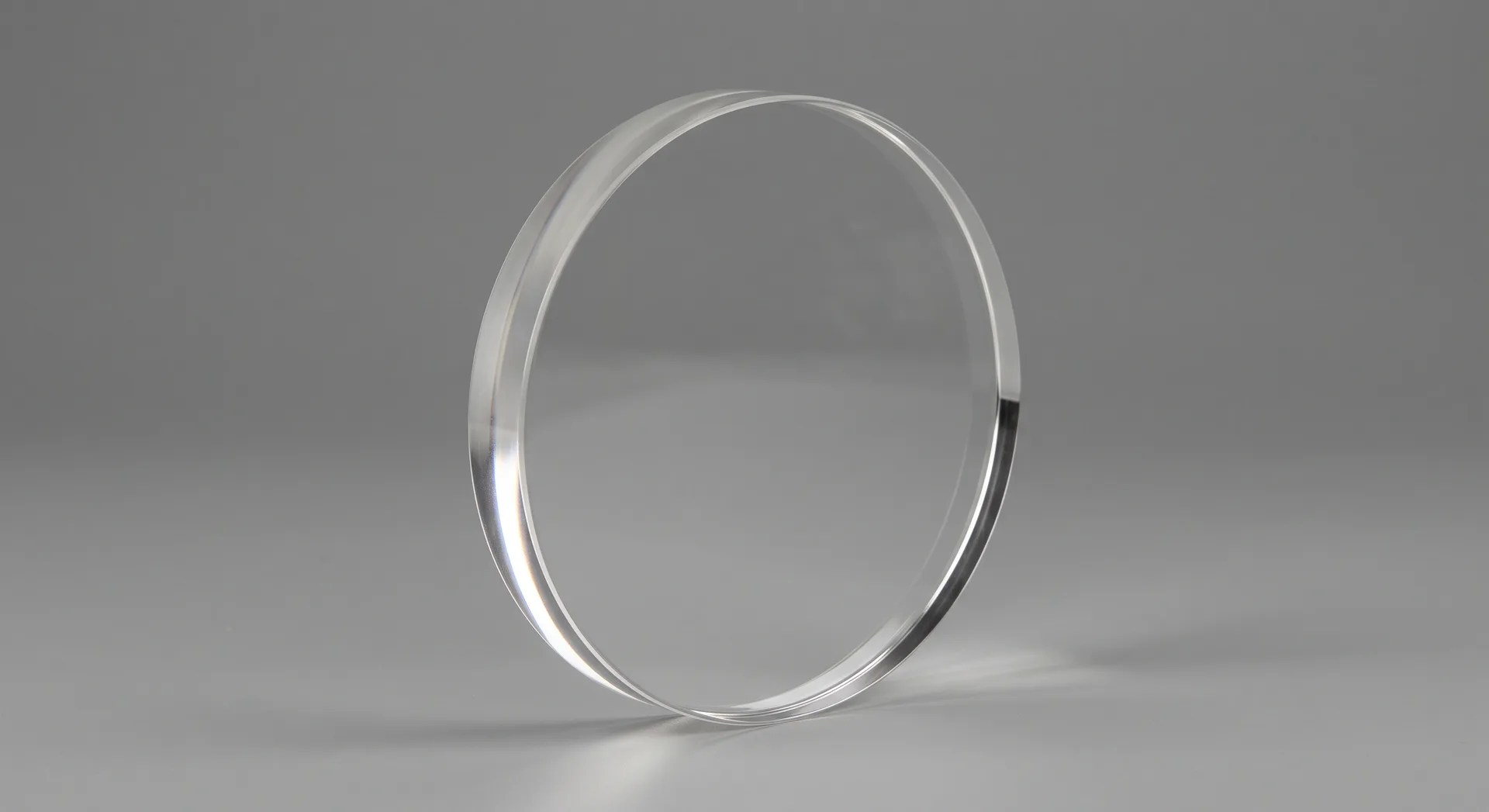 A thick clear ceramic disc standing on a grey surface, the surface behind it showing through softened, a bright line of light along one edge.